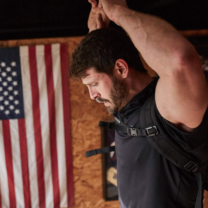 A man in a black shirt stretches his arms overhead in a gym, with the US flag hanging on the wooden wall. Nearby, GORUCK's Training Sternum Strap with MOLLE webbing and adjustable weight distribution suggests intense training sessions that await.