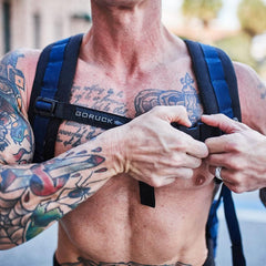 A shirtless, tattooed individual adjusts the GORUCK Training Sternum Strap on their blue backpack with MOLLE webbing, ensuring perfect weight distribution while outdoors. The image showcases their inked chest and arms as they prepare for an adventure.