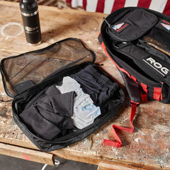 Open black packing cube with folded clothes on rustic wooden table next to black GORUCK water bottle and packing bag