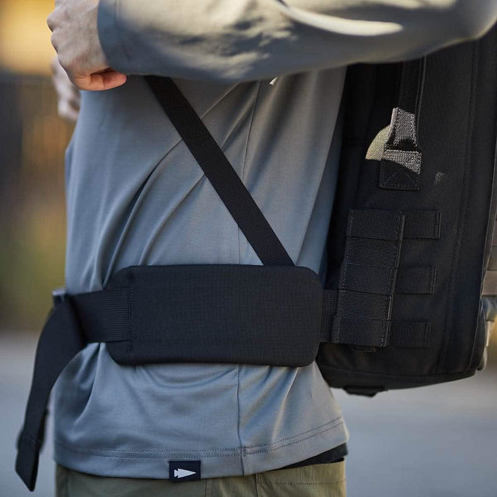 A person sporting a gray long-sleeve shirt adjusts the black strap of their GORUCK backpack. This backpack, featuring various compartments and a Padded Hip Belt ideal for carrying heavy loads, is captured from the side as they stand outdoors against a backdrop of blurred greenery.
