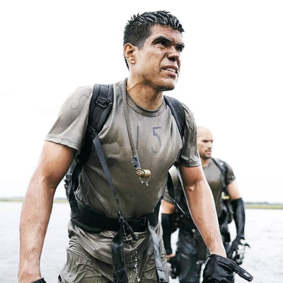 Two men, sporting GORUCK backpacks equipped with Padded Hip Belts and wearing wet uniforms, wade through shallow water with determination and focus. They seem engaged in a challenging outdoor activity or training exercise, likely bearing heavy loads. An overcast sky serves as the backdrop.