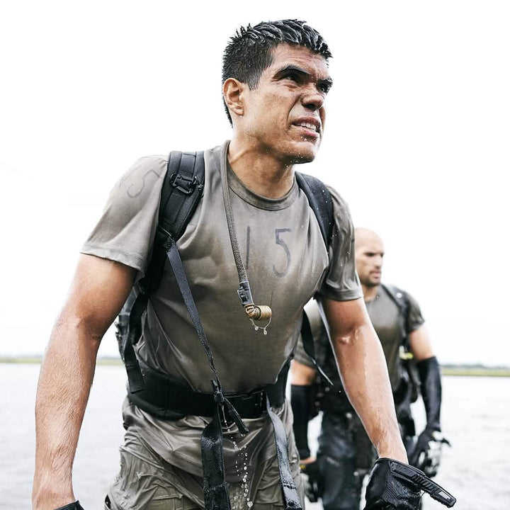 Two men, sporting GORUCK backpacks equipped with Padded Hip Belts and wearing wet uniforms, wade through shallow water with determination and focus. They seem engaged in a challenging outdoor activity or training exercise, likely bearing heavy loads. An overcast sky serves as the backdrop.