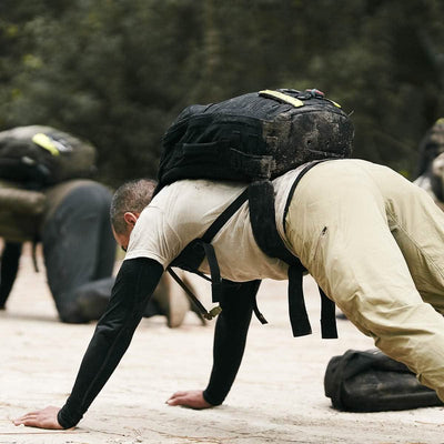 A man in outdoor athletic gear performs a bear crawl on a dirt surface, wearing a backpack equipped with the GORUCK Padded Hip Belt. Others dressed similarly are visible in the background, participating in physical activities. The scene is set in a wooded area designed for training with heavy loads.
