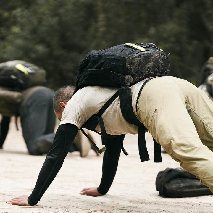 A man in outdoor athletic gear performs a bear crawl on a dirt surface, wearing a backpack equipped with the GORUCK Padded Hip Belt. Others dressed similarly are visible in the background, participating in physical activities. The scene is set in a wooded area designed for training with heavy loads.