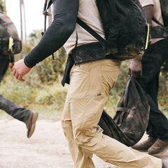Individuals sporting GORUCK backpacks equipped with a Padded Hip Belt and beige pants traverse a dirt path in an outdoor environment. The image showcases a side perspective of their legs and portions of the backpacks, tailored for heavy loads. The backdrop reveals lush greenery and a softly blurred natural setting.