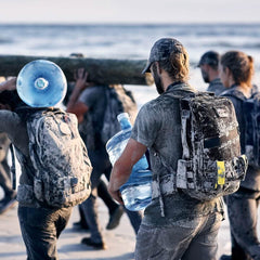The group, dressed in outdoor gear and surrounded by sand, exercises intensely on the beach. One individual carries a large water jug secured with a GORUCK Padded Hip Belt, while others heft substantial loads such as a massive log. The scene is vibrant and implies teamwork and endurance in this coastal setting.