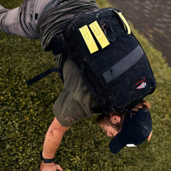A person wearing an army green shirt and cap is doing a push-up on grass, equipped with a GORUCK Padded Hip Belt backpack in black, accented with yellow highlights for carrying heavy loads. The bag includes a patch with red, white, and blue stitching. A body of water is partially visible in the background.