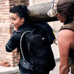 Two people carry a large wooden log on their shoulders. The woman in the foreground, equipped with a black vest and GORUCK backpack featuring a hydration tube, benefits from the comfort of a Padded Hip Belt. The person next to her wears a camo cap as they both appear focused and determined.