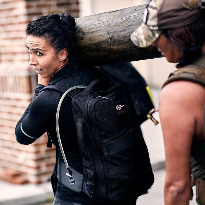 Two people carry a large wooden log on their shoulders. The woman in the foreground, equipped with a black vest and GORUCK backpack featuring a hydration tube, benefits from the comfort of a Padded Hip Belt. The person next to her wears a camo cap as they both appear focused and determined.
