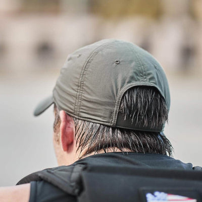 A person sporting the GORUCK Performance TAC Hat - TOUGHDRY in olive green, featuring a VELCRO® closure, is captured from behind. Their hair looks wet beneath the cap made from ToughDry® fabric, as they wear a black top against a blurred background.