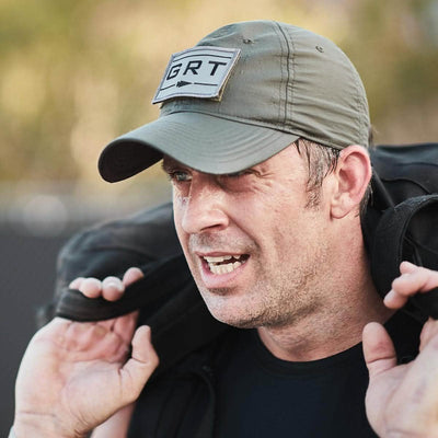 A man wearing a green Performance TAC Hat by GORUCK, featuring a GRT patch, carries a heavy backpack. The cap's ToughDry® fabric and sweat-wicking technology suggest he's well-prepared for the exertion of outdoor adventures, set against a blurred natural background.
