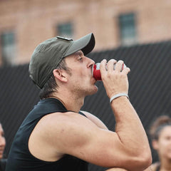 A man in a GORUCK Performance TAC Hat - TOUGHDRY sips from a red can outdoors while wearing a black sleeveless shirt. He sits with others against the backdrop of a blurred brick building and black fence.