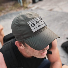 A person wearing an olive green Performance TAC Hat - TOUGHDRY from GORUCK, featuring a GRT patch, sits outdoors. The hat's ToughDry fabric matches their black sleeveless shirt as they slightly crouch on a concrete surface. A backpack is partially visible in the background, ready for adventure.