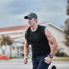 A man in a black sweat-wicking tank top and a GORUCK Performance TAC Hat - TOUGHDRY runs outside, wearing a backpack and a smartwatch. The background reveals a building and trees under a cloudy sky.