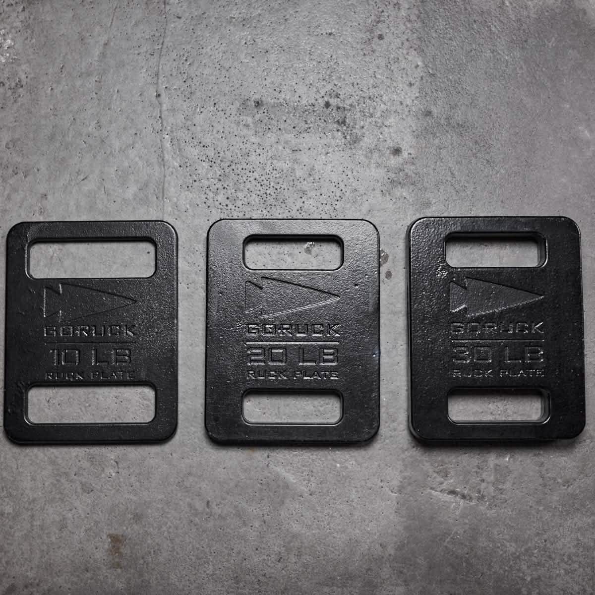 Three GORUCK Ruck Plates are lined up on a concrete surface, exemplifying superior American manufacturing. Each black plate, crafted for the Rucker, is labeled with weights of 10 LB, 20 LB, and 30 LB and includes cut-out handles at both the top and bottom for convenient handling.