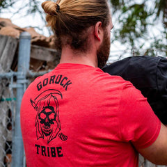 Man with long hair outdoors near a fence carries a black object while wearing the red "Life Has Risk" T-shirt, reflecting his connection to the GORUCK Tribe community.