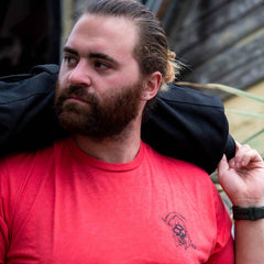 Bearded man in a "Life Has Risk" Red T-shirt from GORUCK, carrying a black shoulder bag and looking to the side, embodies the GORUCK Tribe spirit and community.