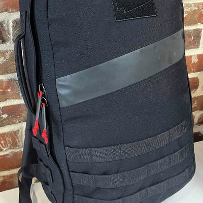 A GORUCK Paracord Kit - Built By SCARS backpack featuring a black design with a diagonal gray stripe, paracord zipper pulls enhanced by heat shrink tubes, and multiple strap loops is set against a brick wall background.