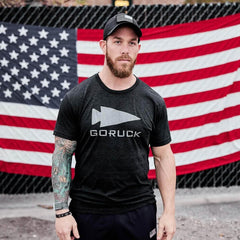 Man in GORUCK reflective tee and cap stands in front of large American flag outdoors