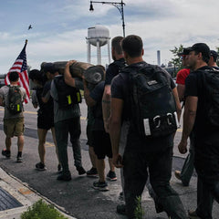 Group of people rucking with backpacks while carrying a log on a street near a water tower and American flag