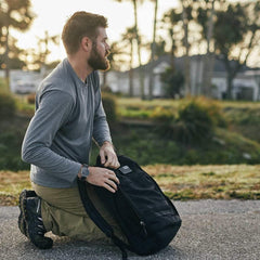 Bearded man in outdoor setting kneeling while holding a black GORUCK rucksack on pavement