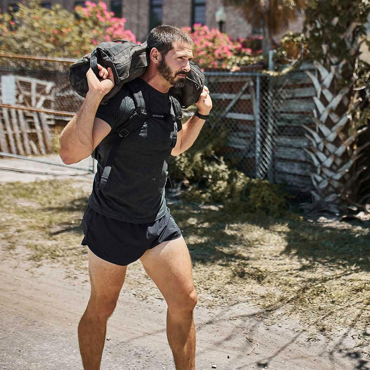 A man with a beard and short hair carries a weighted backpack on his shoulders while walking outdoors, sporting GORUCK's Patch - Ranger Panties Ruck. He wears a black t-shirt, an ensemble typical for a ruck club. In the background, there's a fence, lush trees, and an imposing building.