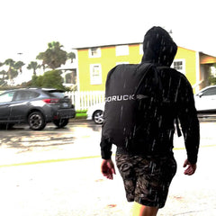 A person in a black hoodie and camouflage shorts walks in the rain, carrying a GORUCK waterproof ruck rain cover. Cars and a yellow building provide a backdrop.