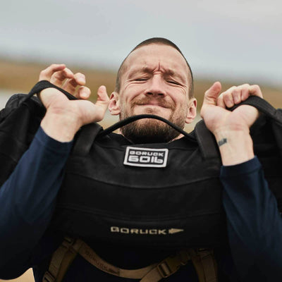 A person with a strained facial expression is lifting a heavy GORUCK Sandbags 2.0 while wearing a long-sleeve shirt, set against blurred fields in the background. The "Scars Lifetime Guarantee" logo is prominently displayed on the 1000D CORDURA Sandbags 2.0.