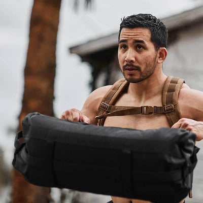 A man with a focused expression is lifting the heavy black GORUCK Sandbags 2.0 outdoors, highlighting the durability of 1000D CORDURA. He wears a brown harness and has short dark hair. A blurred palm tree and building hint at his intense training session backed by Scars Lifetime Guarantee.