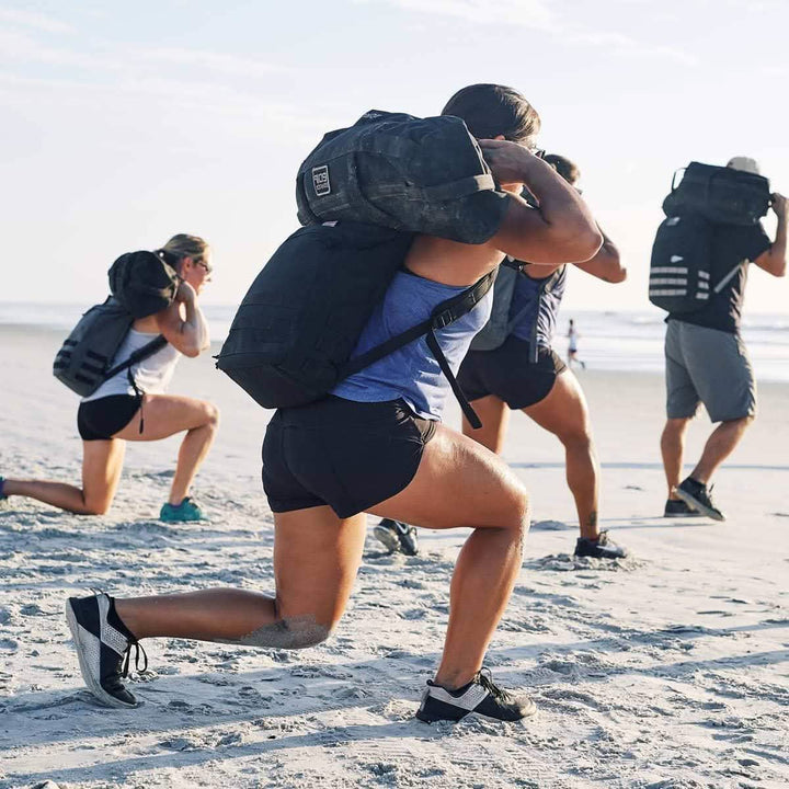 Exercise enthusiasts on a beach are performing lunges with their Patch - Sandbag Weights from vendor-unknown, as the sun glistens over the ocean. The group, a mix of men and women, pushes through their workout on the sandy shore under the clear sky.