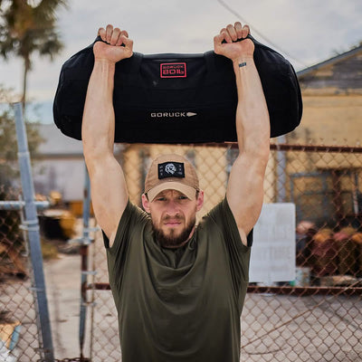 A person wearing a green t-shirt and cap showcases their tactical fitness by hoisting a large black sandbag overhead. They stand outdoors, against the backdrop of a chain-link fence and an aged building, as the cloudy sky enhances the atmosphere of their GORUCK Sandbag Strength with Cadre DS (6-Week Training Program) workout.