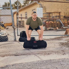 Man in green t-shirt and cap lifting a black GORUCK sandbag in an outdoor urban setting