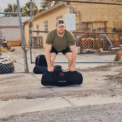 A man in a green t-shirt and black shorts demonstrates his prowess with a GORUCK sandbag during the Sandbag Strength with Cadre DS (6-Week Training Program) outdoors, near a chain-link fence and a beige building. Other bags and wooden pallets litter the concrete ground, offering an ideal scene for tactical fitness.