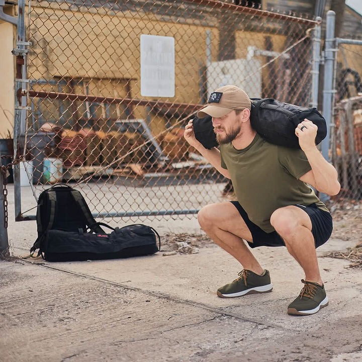 A man in a green shirt and black shorts demonstrates a squat from the GORUCK Sandbag Strength with Cadre DS, emphasizing tactical fitness. He is working out outdoors on a sidewalk next to a chain-link fence and industrial background, with his backpack placed nearby.