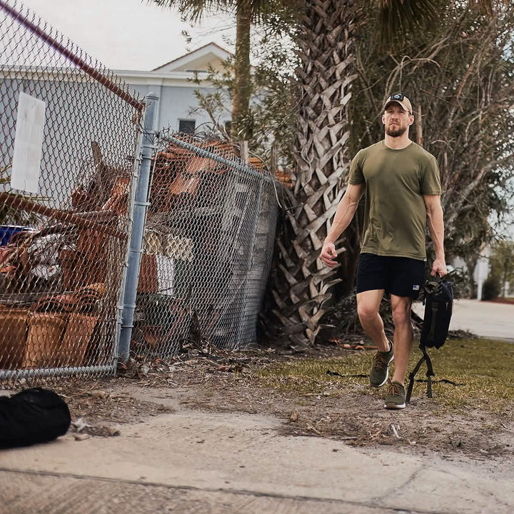 A man in gear supporting the Sandbag Strength with Cadre DS 6-Week Training Program by GORUCK demonstrates tactical fitness as he walks alongside a chain-link fence, carrying a black backpack. With trees in the background, he is wearing an olive green t-shirt, black shorts, a cap, and sneakers.
