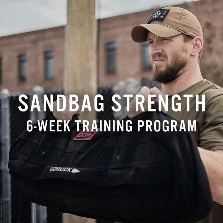 A man sporting a cap hoists a hefty sandbag outdoors, exemplifying tactical fitness. The image text reads, Sandbag Strength with Cadre DS (6-Week Training Program) by GORUCK. In the backdrop, a chain-link fence and brick building create the gritty atmosphere for ultimate SALT conditioning.