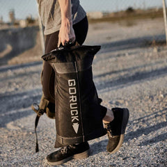 Dressed in athletic gear, an individual walks along a gravel path carrying a black Simple Training Sandbag by GORUCK. The long shadows cast by the sun accentuate their legs in black sneakers. In the distance, there's a chain-link fence against a blurred background, echoing the durability assured by GORUCK's Scars Lifetime Guarantee.