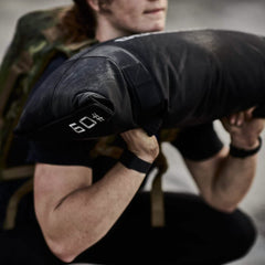 A person dressed in athletic wear is lifting a GORUCK Simple Training Sandbag marked with 69. They are outdoors and appear focused on their exercise. Wearing a wristband and carrying a backpack, they demonstrate durability similar to Scars Lifetime Guarantee.