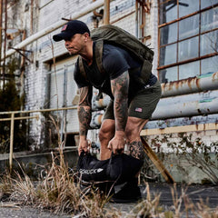 A man wearing a dark cap and shorts, showcasing tattoos on his arms and legs, is training with a black GORUCK Simple Training Sandbag outdoors near an industrial building. Carrying a backpack, he embodies the grit of outdoor workouts, counting on the Scars Lifetime Guarantee as he moves through the grass.