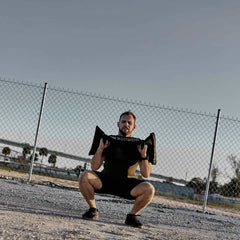 A man is performing outdoor training with a GORUCK Simple Training Sandbag near a chain-link fence. He is squatting while holding the sandbag at shoulder height under sunny skies, with a gravelly ground and clear weather, creating an ideal scene for fitness enthusiasts seeking durability and performance.