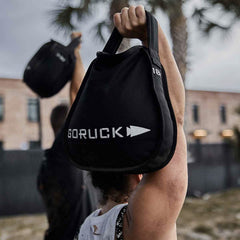 With a backdrop of a brick building and palm trees, an individual hoists a black GORUCK Sand Kettlebell overhead outdoors, highlighting the product's endurance-focused design as a blurred figure mirrors the action, capturing the essence of resilience and strength.