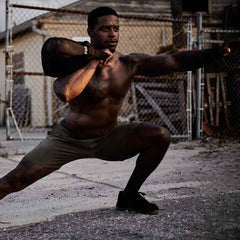 A man is engaged in an outdoor workout, lunging with a GORUCK Sand Kettlebell on his shoulder. He's shirtless and dressed in shorts and sneakers, with a chain-link fence and building set against soft lighting in the background.