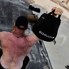 A sunburned individual, without a shirt, is using one arm to lift a GORUCK Sand Kettlebell and is donning a black cap while seated on a textured surface that highlights its endurance design. In the background, two sandbags can be seen.