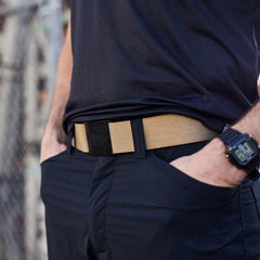 Close-up of a person wearing a tan tactical belt with a sturdy black buckle and navy pants, hands in pockets