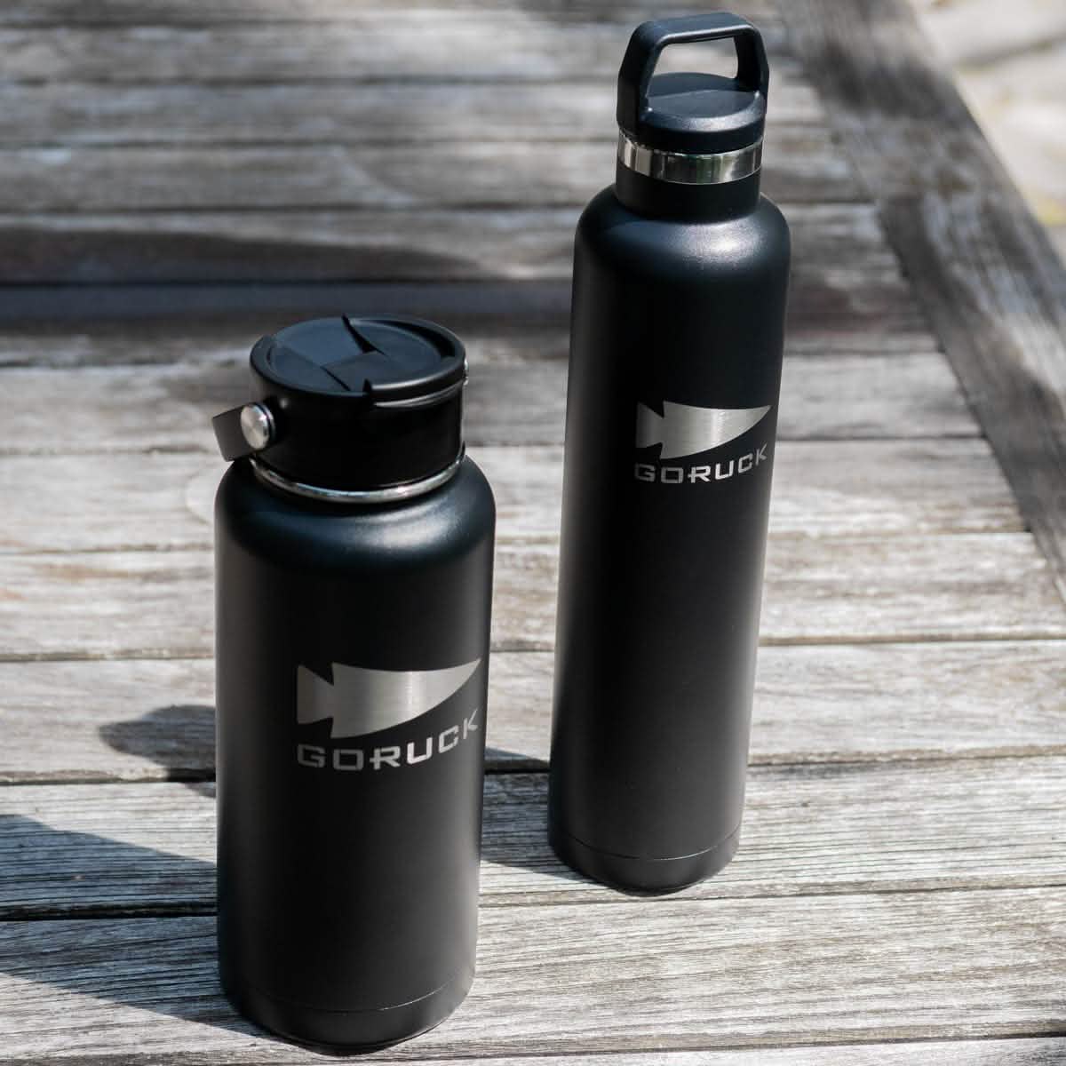 Two black metal GORUCK x RTIC water bottles, crafted from stainless steel, stand on a wooden surface. One bottle features a flip-top lid, while the other has a screw-top lid with a handle. Their double wall insulated design ensures beverages stay at the right temperature on the naturally weathered table.