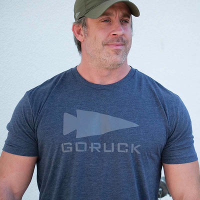 A semi-famous man in a premium Three Rules Tee - Tri-Blend, dark blue with an arrowhead design, stands before a light-colored wall. Sporting a light beard and green baseball cap, he gazes slightly to the side with casual confidence.