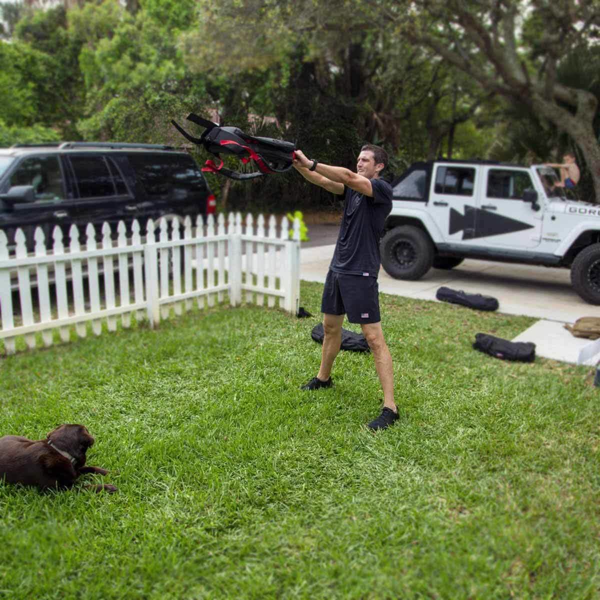 A person in GORUCK's Men’s USA Training Shorts - ToughStretch exercises outdoors with a kettlebell while a brown dog lies on the grass nearby. Two vehicles are parked on the street, and a white picket fence borders the yard. Trees and lush greenery surround the area, creating an ideal backdrop for an active lifestyle.