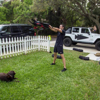 A person in GORUCK's Men’s USA Training Shorts - ToughStretch exercises outdoors with a kettlebell while a brown dog lies on the grass nearby. Two vehicles are parked on the street, and a white picket fence borders the yard. Trees and lush greenery surround the area, creating an ideal backdrop for an active lifestyle.