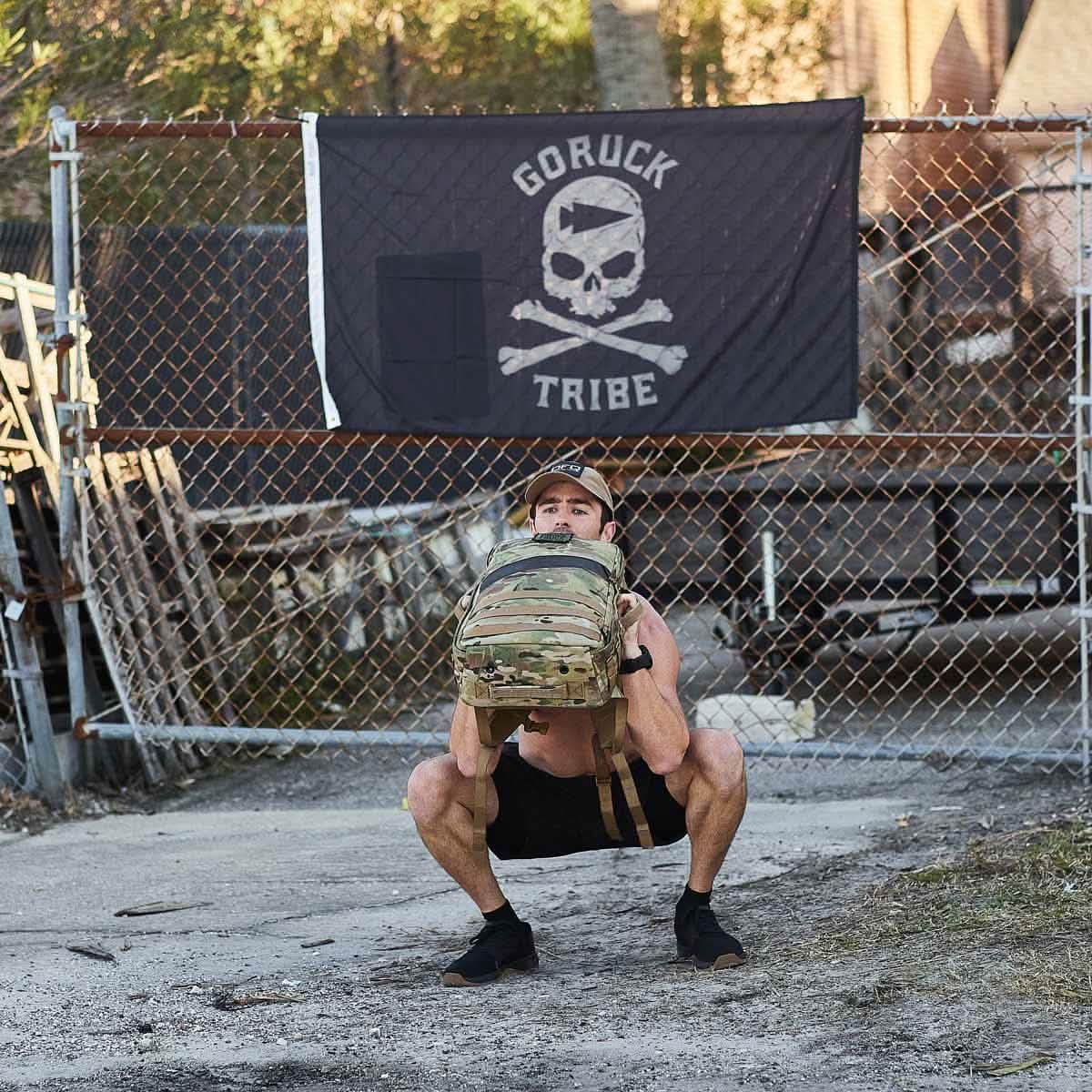 A person squats with a backpack outside, in front of a 
