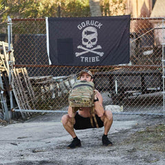 Man doing squats with GORUCK rucksack in front of GORUCK Tribe flag outdoors